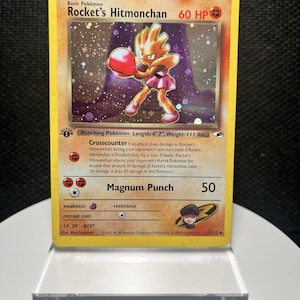 May include: A Rocket's Hitmonchan trading card with a yellow border and a holographic illustration of the character. The card displays 60 HP, the Crosscounter and Magnum Punch attacks, and includes the text Rocket's Hitmonchan, 11/132, and 1995-2000.