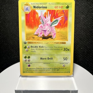 May include: A Nidorino trading card from the 1999 Wizards of the Coast set, featuring a pink creature illustration, 60 HP, and the attacks Double Kick and Horn Drill. The card includes a first edition stamp and text detailing its stats and abilities.