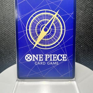 May include: A rectangular One Piece Card Game trading card featuring a deep blue background with a white grid pattern. A large gold compass graphic is centered above the white text that reads ONE PIECE CARD GAME. The card is in a clear case.