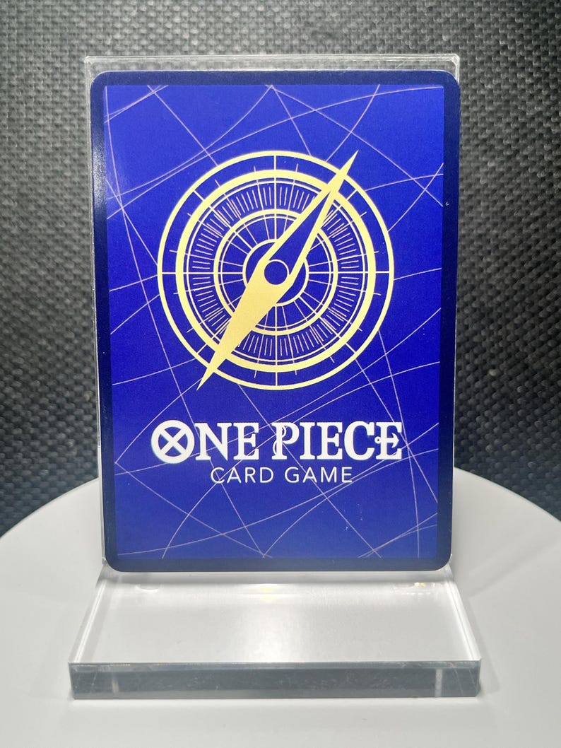 May include: A rectangular One Piece Card Game trading card featuring a deep blue background with a white grid pattern. A large gold compass graphic is centered above the white text that reads ONE PIECE CARD GAME. The card is in a clear case.