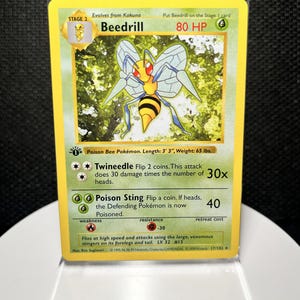 Puede incluir: Una carta coleccionable de Beedrill con un insecto amarillo y negro de ojos rojos y alas. La carta muestra 80 HP, el texto Poison Bee Pokémon, los ataques Twineeedle y Poison Sting, y un sello de 1st Edition sobre un borde amarillo.