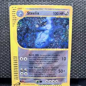May include: A Steelix Pokémon trading card with a holographic blue and silver image of the Steelix character. The card has a yellow border and includes text such as "100 HP" and "Iron Smash".