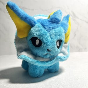 May include: A plush toy featuring a blue and white creature with yellow accents on its ears. The toy has black eyes and a white collar. The plush is made of soft, fluffy material and is standing upright.