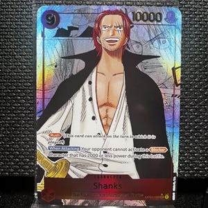 May include: A holographic trading card featuring the character Shanks from the One Piece franchise. The card has a red and black border, with the character's name and card details. The card has a power level of 10000 and includes text describing its abilities.