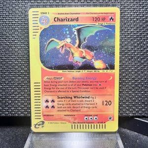 Op de afbeelding: Een Charizard Pokémon-kaart, Fase 2, met een oranje draak met groene vleugels. De kaart heeft een gele achtergrond met tekst die de vaardigheden en statistieken van het personage beschrijft, inclusief 120 HP. De kaart staat op een transparante standaard.