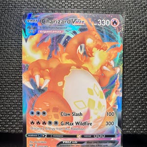 May include: A Charizard VMAX Pokémon card featuring a fiery orange dragon with a white and yellow egg-shaped design. The card has the text "Charizard VMAX" and "Gigantamax" with attack details and a HP of 330.