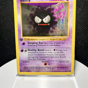 May include: A Gastly trading card with a yellow border and purple background. The card features the character Gastly, 30 HP, and the text 'iowa city gg!'. It lists attacks Sleeping Gas and Destiny Bond, with a 1st Edition stamp and 1999 copyright.