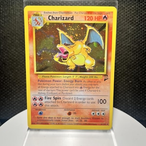 May include: A Charizard trading card featuring a yellow border, a fire-breathing dragon illustration, and 120 HP. The card displays the text Charizard, Flame Pokémon, Energy Burn, and Fire Spin, with a 100 damage value and a 1995-2000 copyright date.