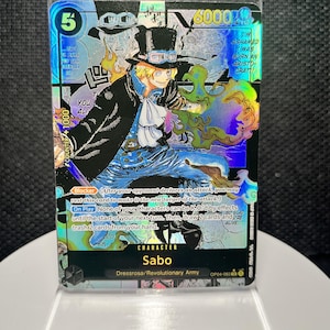 May include: A holographic One Piece trading card featuring the character Sabo in a top hat and goggles, surrounded by green flames. The card displays text including 6000 power, Blocker ability, and On Play effects, set against a manga panel background.