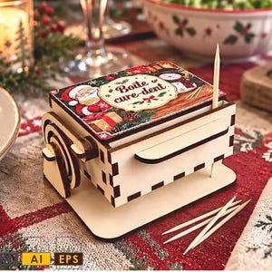Puede incluir: Un dispensador de palillos de madera rectangular con una tapa de temática navideña que muestra ilustraciones de Santa Claus y un muñeco de nieve, con el texto en francés Boîte cure-dent impreso en la parte superior, sobre una base plana.