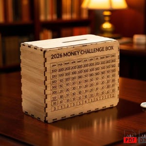 Op de afbeelding: Een houten 2026 Money Challenge Box met een gleuf aan de bovenkant. De doos is gegraveerd met een spaartabel, met bedragen van €3 tot €500. De doos is gemaakt van lichtgekleurd hout en staat op een donker houten oppervlak.