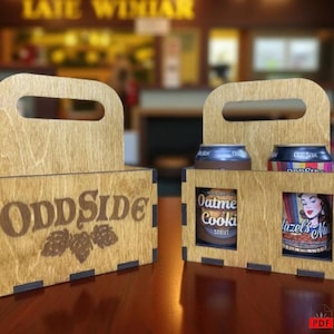 Peut inclure: Deux supports à bière en bois avec poignée, l'un vide et l'autre contenant deux canettes. Les supports portent le texte "OddSide" et un motif de houblon. Les canettes sont étiquetées "Oatmeal Cookie Stout" et "Hazel's Nut".