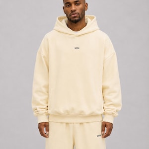 May include: A cream-coloured hooded sweatshirt and matching sweatpants set. The hoodie features a hood, long sleeves with ribbed cuffs, and the text WRN printed in small black letters on the centre chest. The sweatpants have the text WRN in black.