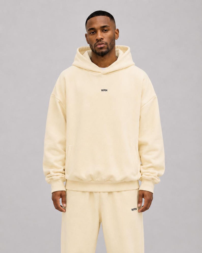 May include: A cream-coloured hooded sweatshirt and matching sweatpants set. The hoodie features a hood, long sleeves with ribbed cuffs, and the text WRN printed in small black letters on the centre chest. The sweatpants have the text WRN in black.