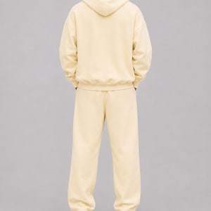 May include: A cream-coloured matching set consisting of a long-sleeved hooded sweatshirt and loose-fitting sweatpants. The hoodie features a hood and ribbed cuffs, while the sweatpants have a straight-leg cut. The outfit is paired with white trainers.