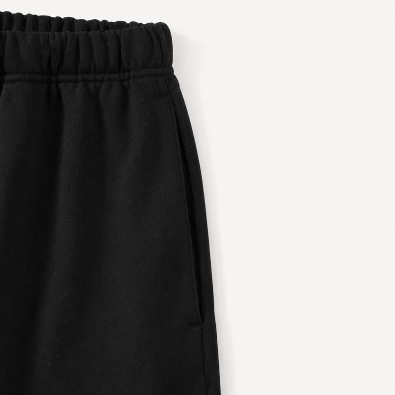 May include: Close-up of black sweatpants with an elasticated waistband and a side pocket. The fabric appears to be a soft, matte material. The trousers are against a white background.