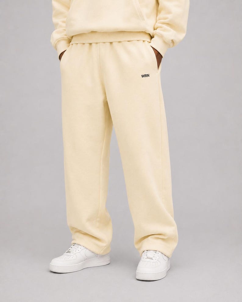 K&ouml;nnte beinhalten: Cremefarbene Jogginghose mit geradem Beinverlauf und elastischem Bund. Im Bereich des linken Oberschenkels befindet sich der schwarze Schriftzug WRN in einer kleinen, serifenlosen Schriftart. Die Hose wird mit wei&szlig;en Sneakern und einem Sweatshirt kombiniert.