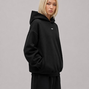 K&ouml;nnte beinhalten: Ein schwarzer Kapuzenpullover ohne Kordelzug mit langen &Auml;rmeln und einem kleinen wei&szlig;en Logo auf der oberen Brust. Das Kleidungsst&uuml;ck hat eine lockere Passform mit gerippten B&uuml;ndchen am &Auml;rmel und am Saum und besteht aus schwarzem Stoff.
