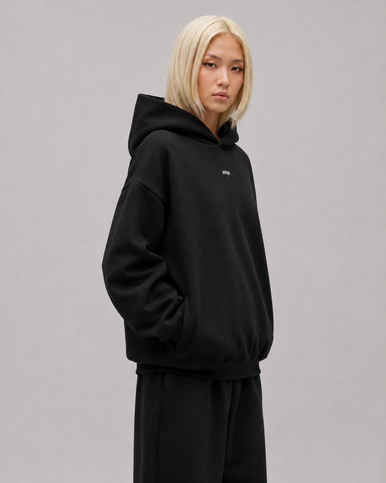 K&ouml;nnte beinhalten: Ein schwarzer Kapuzenpullover ohne Kordelzug mit langen &Auml;rmeln und einem kleinen wei&szlig;en Logo auf der oberen Brust. Das Kleidungsst&uuml;ck hat eine lockere Passform mit gerippten B&uuml;ndchen am &Auml;rmel und am Saum und besteht aus schwarzem Stoff.