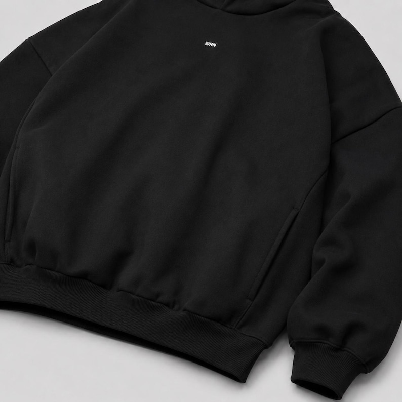 K&ouml;nnte beinhalten: Ein schwarzer Kapuzenpullover mit langen &Auml;rmeln und einem kleinen wei&szlig;en Textlogo mit der Aufschrift WRN, das mittig im oberen Brustbereich platziert ist. Das Kleidungsst&uuml;ck hat einen gerippten Saum und gerippte &Auml;rmelb&uuml;ndchen aus glattem Stoff.