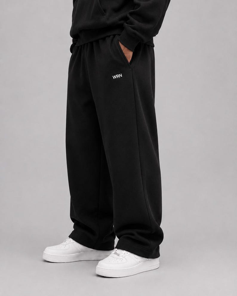 Puede incluir: Pantalones de ch&aacute;ndal negros con silueta de pierna ancha y cintura el&aacute;stica. El texto WRN est&aacute; impreso en blanco en la parte superior del muslo izquierdo. Estos pantalones se combinan con zapatillas blancas, creando un estilo casual y s&oacute;lido.