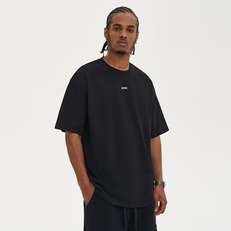 May include: Black short-sleeved t-shirt with the white text "WRN" centered on the chest. The shirt has a relaxed fit and is worn by a person. The person is wearing a watch on their left wrist.