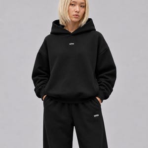 K&ouml;nnte beinhalten: Ein schwarzer Kapuzenpullover und eine passende schwarze Jogginghose. Der Kapuzenpullover hat ein kleines wei&szlig;es Textlogo mit der Aufschrift WRN, das mittig auf der Brust platziert ist. Die Hose zeigt dasselbe wei&szlig;e WRN-Logo am linken Oberschenkel.