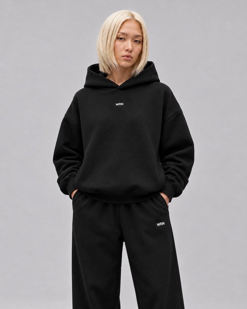 K&ouml;nnte beinhalten: Ein schwarzer Kapuzenpullover und eine passende schwarze Jogginghose. Der Kapuzenpullover hat ein kleines wei&szlig;es Textlogo mit der Aufschrift WRN, das mittig auf der Brust platziert ist. Die Hose zeigt dasselbe wei&szlig;e WRN-Logo am linken Oberschenkel.