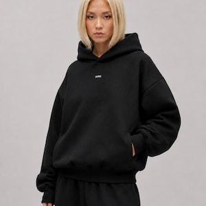 K&ouml;nnte beinhalten: Ein schwarzer Kapuzenpullover mit langen &Auml;rmeln, einer K&auml;ngurutasche auf der Vorderseite und dem Text WRN in kleinen wei&szlig;en Buchstaben mittig auf der Brust. Das Kleidungsst&uuml;ck hat eine lockere Passform mit gerippten B&uuml;ndchen und einem gerippten Saum.