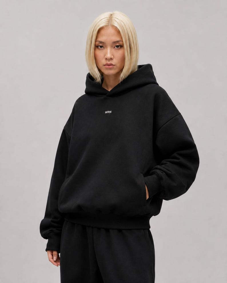 K&ouml;nnte beinhalten: Ein schwarzer Kapuzenpullover mit langen &Auml;rmeln, einer K&auml;ngurutasche auf der Vorderseite und dem Text WRN in kleinen wei&szlig;en Buchstaben mittig auf der Brust. Das Kleidungsst&uuml;ck hat eine lockere Passform mit gerippten B&uuml;ndchen und einem gerippten Saum.