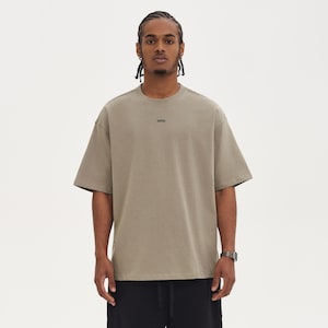 May include: A light brown, oversized t-shirt with short sleeves. The word "WRN" is printed in black on the chest. The person in the image is wearing black shorts and a silver watch.
