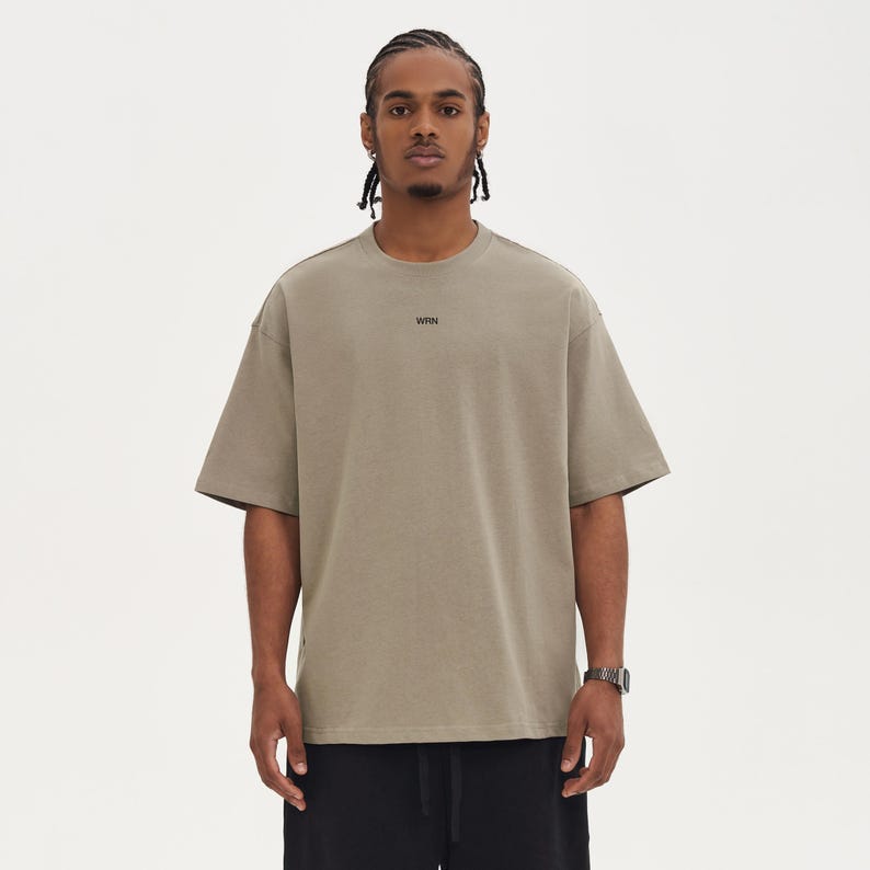 May include: A light brown, oversized t-shirt with short sleeves. The word "WRN" is printed in black on the chest. The person in the image is wearing black shorts and a silver watch.