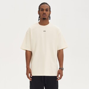 May include: Cream-colored oversized t-shirt with short sleeves. The shirt has the letters "WRN" printed in black on the chest. The person is wearing a silver watch and black pants. The background is plain white.