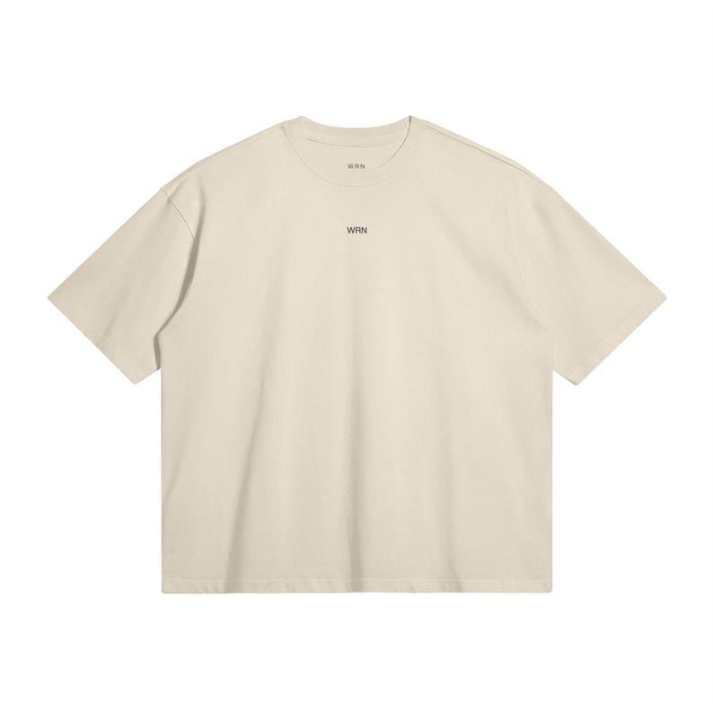 May include: Cream-colored short-sleeved t-shirt with the text "WRN" printed in black near the neckline. The shirt has a relaxed fit and a simple, minimalist design. The fabric appears to be a soft, breathable material.