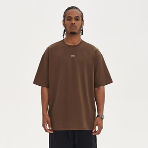 May include: A brown, short-sleeved t-shirt with the word "WRN" printed in white on the chest. The shirt has a relaxed fit and a crew neck. The model is wearing black pants. The background is white.