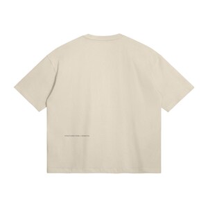 May include: Cream-colored, oversized t-shirt. The back of the shirt features the text "STRUCTURED FORM / ESSENTIAL" printed in a small, dark font near the bottom hem. The shirt has short sleeves and a crew neck.