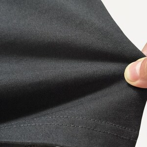 May include: Close-up of a dark gray fabric with a ribbed texture. The fabric is being held, revealing its flexibility and the detailed stitching along the hem. The material appears to be a knit or similar textile.