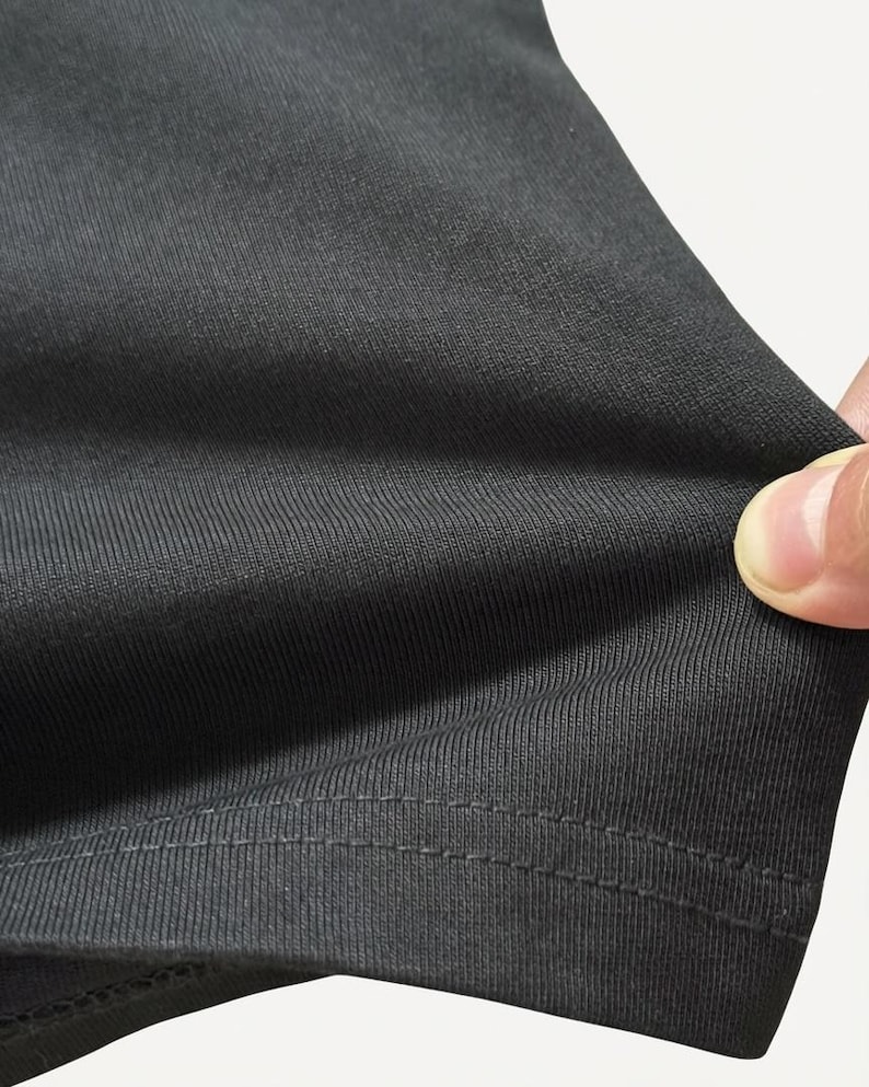May include: Close-up of a dark gray fabric with a ribbed texture. The fabric is being held, revealing its flexibility and the detailed stitching along the hem. The material appears to be a knit or similar textile.
