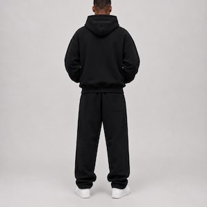K&ouml;nnte beinhalten: Ein einfarbiger schwarzer Kapuzenpullover mit langen &Auml;rmeln und dazu passender schwarzer Jogginghose. Das Outfit hat eine lockere Passform, eine Kapuze sowie gerippte B&uuml;ndchen an &Auml;rmeln und Hosenbeinen. Wei&szlig;e Sneaker vervollst&auml;ndigen das Ensemble.