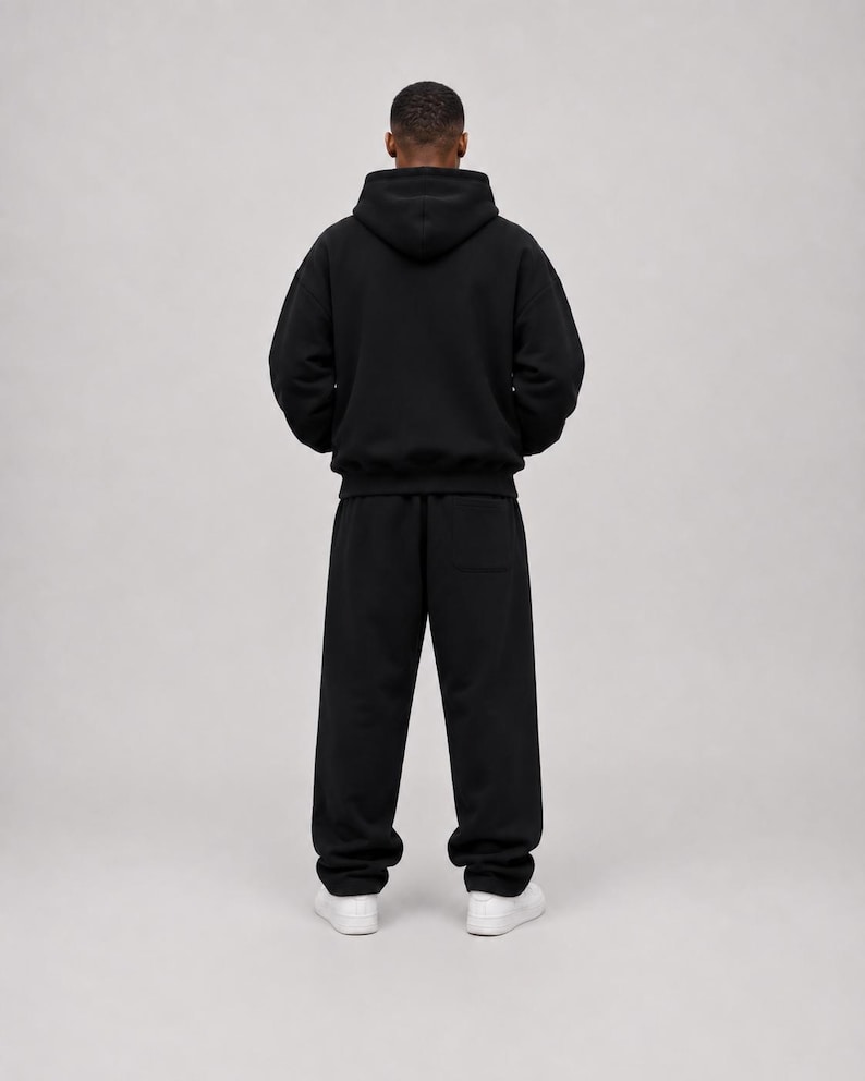 K&ouml;nnte beinhalten: Ein einfarbiger schwarzer Kapuzenpullover mit langen &Auml;rmeln und dazu passender schwarzer Jogginghose. Das Outfit hat eine lockere Passform, eine Kapuze sowie gerippte B&uuml;ndchen an &Auml;rmeln und Hosenbeinen. Wei&szlig;e Sneaker vervollst&auml;ndigen das Ensemble.
