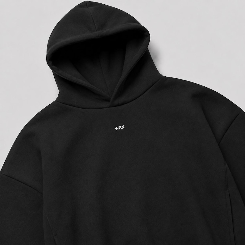 K&ouml;nnte beinhalten: Ein einfarbiger schwarzer Kapuzenpullover mit einer Kapuze ohne Kordelzug und langen &Auml;rmeln. In der Mitte der Brust befinden sich die Buchstaben WRN, die in einer kleinen, wei&szlig;en, serifenlosen Schrift aufgedruckt sind. Der Stoff ist glatt und matt.