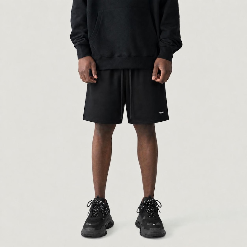 May include: Black shorts with a drawstring waist and a small white logo. The shorts are paired with a black hoodie and black trainers with patterned laces. The outfit is casual and sporty.