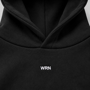 K&ouml;nnte beinhalten: Ein einfarbiger schwarzer Kapuzenpullover mit einer Kapuze ohne Kordelzug und dem wei&szlig;en Schriftzug WRN auf der Brust, der in einer klaren serifenlosen Schrift gedruckt ist. Das Kleidungsst&uuml;ck besteht aus glattem, mattem Stoff.