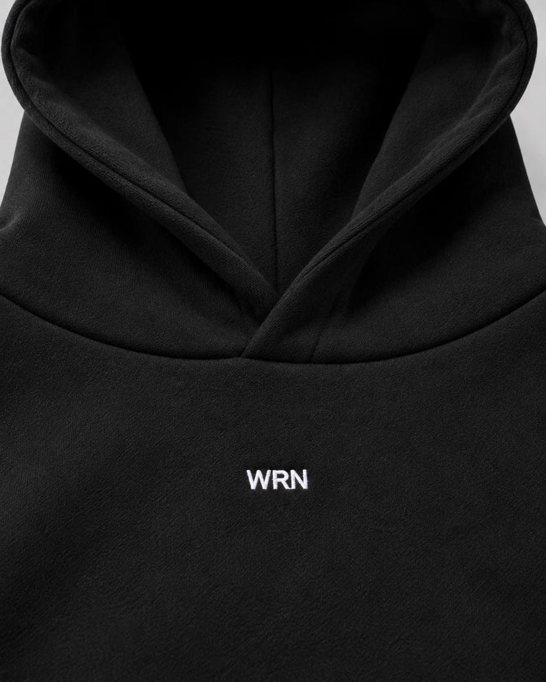K&ouml;nnte beinhalten: Ein einfarbiger schwarzer Kapuzenpullover mit einer Kapuze ohne Kordelzug und dem wei&szlig;en Schriftzug WRN auf der Brust, der in einer klaren serifenlosen Schrift gedruckt ist. Das Kleidungsst&uuml;ck besteht aus glattem, mattem Stoff.