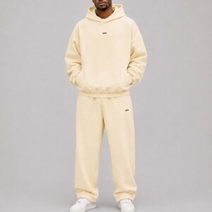 May include: A cream-coloured matching set consisting of a long-sleeve hooded sweatshirt and loose-fitting sweatpants. The hoodie features a small black text logo on the centre chest, and the trousers have a matching logo on the left thigh.