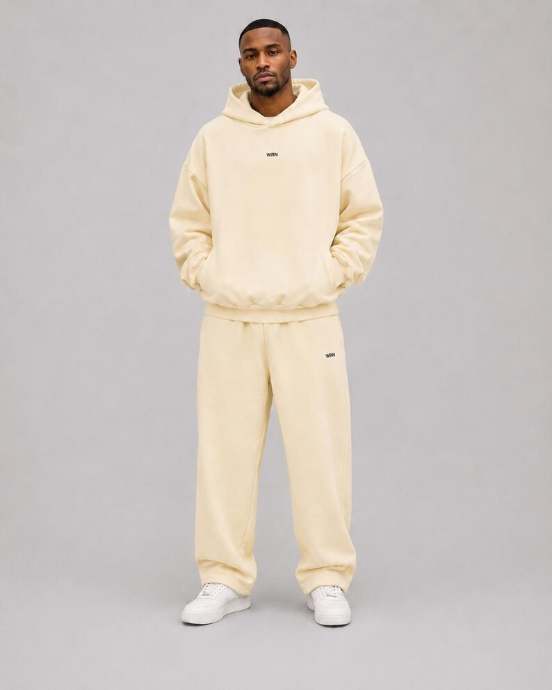 May include: A cream-coloured matching set consisting of a long-sleeve hooded sweatshirt and loose-fitting sweatpants. The hoodie features a small black text logo on the centre chest, and the trousers have a matching logo on the left thigh.
