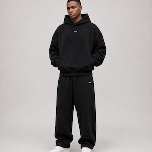 K&ouml;nnte beinhalten: Ein schwarzer Kapuzenpullover und eine dazu passende schwarze Jogginghose. Der Hoodie hat ein kleines wei&szlig;es Logo auf der Brust und die Jogginghose ein kleines wei&szlig;es Logo auf dem linken Bein. Das Outfit wird mit wei&szlig;en Sneakern kombiniert.
