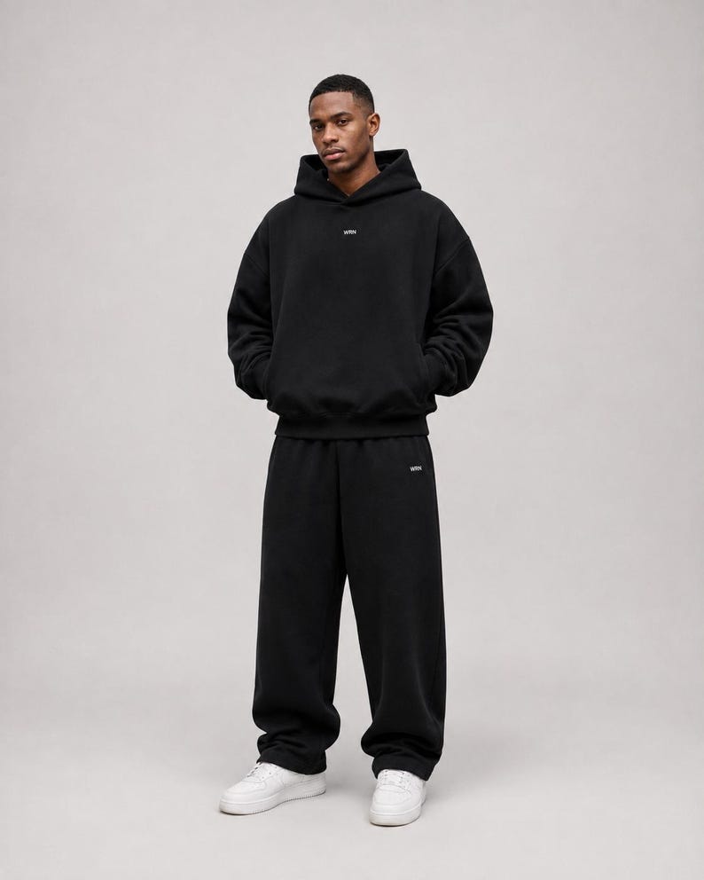 K&ouml;nnte beinhalten: Ein schwarzer Kapuzenpullover und eine dazu passende schwarze Jogginghose. Der Hoodie hat ein kleines wei&szlig;es Logo auf der Brust und die Jogginghose ein kleines wei&szlig;es Logo auf dem linken Bein. Das Outfit wird mit wei&szlig;en Sneakern kombiniert.