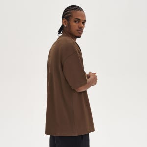 May include: A brown short-sleeve t-shirt with a relaxed fit. The shirt has a crew neck and a slightly oversized silhouette. The fabric appears to be a soft, solid color material. The person is wearing black pants.