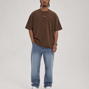 May include: A person wearing a brown t-shirt with a small logo and blue jeans. The t-shirt is a solid color and has short sleeves. The jeans are a light blue wash and have a relaxed fit. White sneakers complete the outfit.